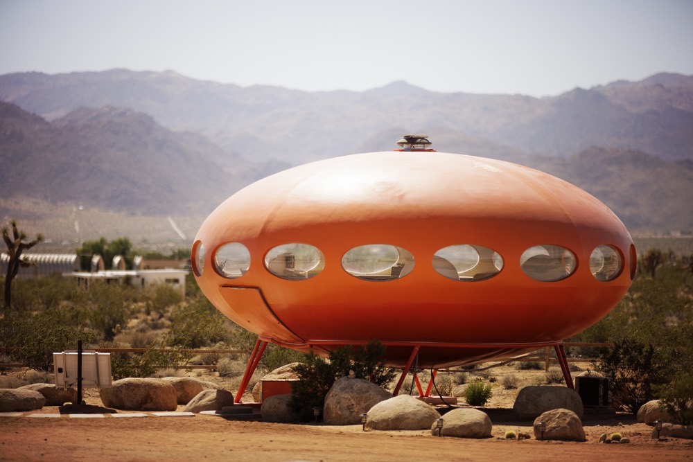 A round orange Futuro house on stilts in front of desert mountains